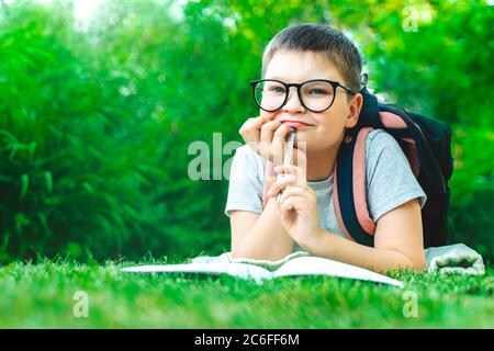 Glücklich Schuljunge in Brille Studium im grünen Park Blick auf Kamera schreiben im Buch Hausaufgaben. Lächelndes Kind macht Notizen im Freien im Park. Zurück t Stockfoto