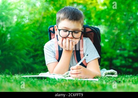 Ernst niedlichen Schuljunge in Brille mit Tasche Pack lag auf Gras schreiben Hausaufgaben im grünen Garten. Männlich Kind Zeichnung Notizen machen Mathe im Freien. Stockfoto