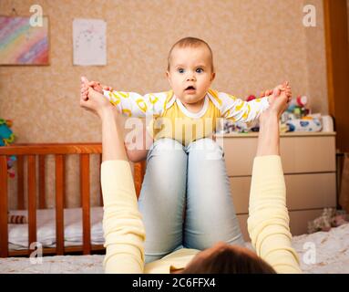 Fürsorgliche unbekannte junge Mutter in lässiger Kleidung spielt Flugzeug mit ihrem charmanten halbjährigen hübschen Tochter auf dem Bett im Kinderzimmer. Mothe Stockfoto