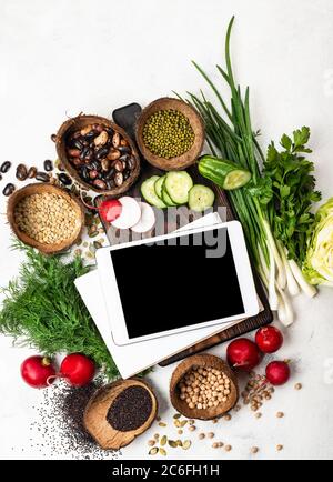 Bestellen Sie Lebensmittel online Konzept. Tablet-Computer mit leerem Bildschirm und Notizblock auf einem Schneidebrett mit Zutaten zum Kochen vegane Lebensmittel Draufsicht. Stockfoto