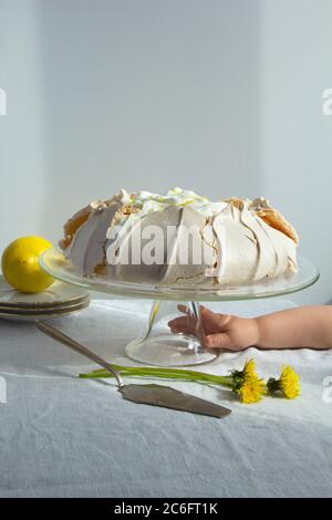 Hausgemachte Baiser-Kuchen Pavlova mit Schlagsahne auf Glas Kuchen Stand. Kind der Hand greift nach dem Kuchen auf dem Tisch. Knuspriger Nachtisch auf dem weißen Backgro Stockfoto