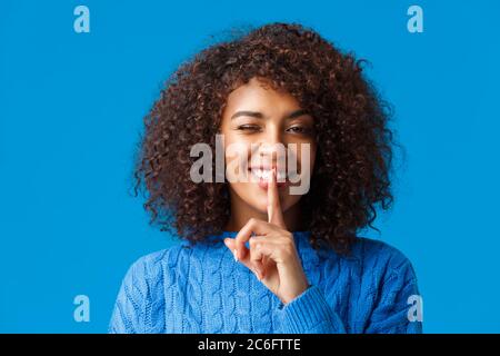 Unbeschwerte fröhliche afro-amerikanische Hüftmädchen, mit stylischem Afro-Haarschnitt, kühlem und sassy winken, lächelnd Shush-Geste machen, Zeigefinger auf die Lippen drücken Stockfoto