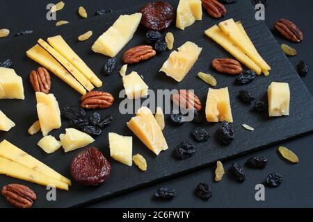 Stücke von Parmesan und gouda mit Bockshornklee, blauen und goldenen Rosinen und getrockneten Aprikosen auf schwarzem Steinteller auf schwarzem Tisch. Nahaufnahme Stockfoto