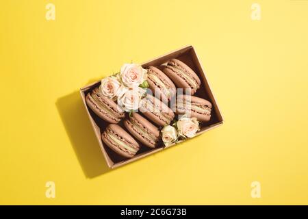 Flache Lay von vielen Schokolade französisch Dessert Macarons in Handwerk Box mit schönen beigen Rosen auf dem gelben Hintergrund. Stockfoto
