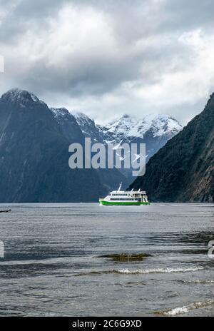 Touristenboot fährt durch Milford Sound, hinter Bergspitzen mit Schnee, Southland, South Island, Neuseeland Stockfoto