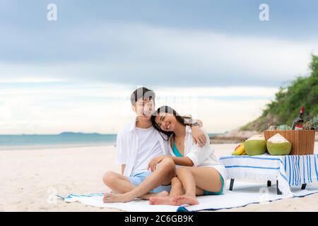 Asiatisches junges Paar sitzt auf der Picknickdecke und entspannen Sie sich am weißen Sandstrand und in der Nähe des Meeres mit tropischen Früchten im Hintergrund. Sommer, Urlaub, V Stockfoto