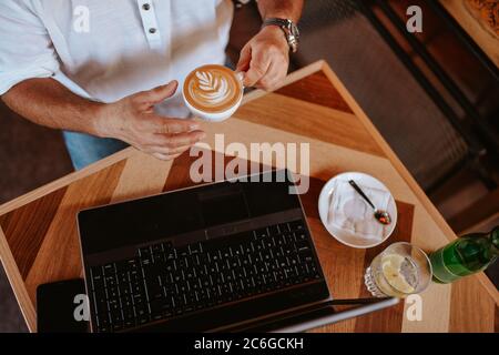 Draufsicht auf die Hände eines kaukasischen Geschäftsmannes mittleren Alters in einem weißen Hemd, der in einem Café sitzt, an einem Laptop arbeitet und Espresso trinkt Stockfoto