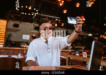 Ein kaukasischer Geschäftsmann mittleren Alters mit Brille in einem weißen Hemd sitzt in einem Café und macht ein Selfie, während ein Laptop auf dem Tisch steht Stockfoto
