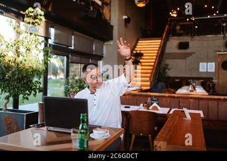 Ein moderner Geschäftsmann mittleren Alters mit Brille in weißem Hemd sitzt in einem Café, arbeitet an einem Laptop und winkt mit der Hand Stockfoto