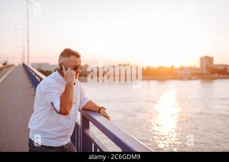 Ein kaukasischer Geschäftsmann mittleren Alters mit Brille, in einem weißen Hemd, und am Telefon gelehnt an das Brückengeländer Stockfoto