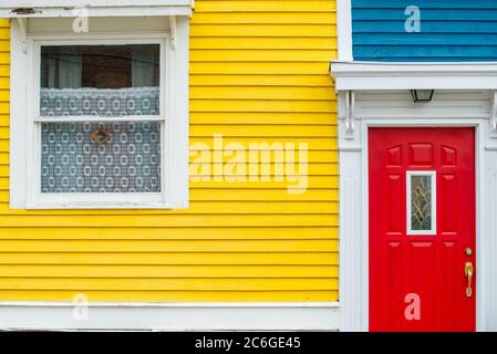 Leuchtend rote Tür eines Gebäudes mit blauen und bunten gelben Holztäfelwänden. Die Metalltür hat ein kleines Fenster. Stockfoto