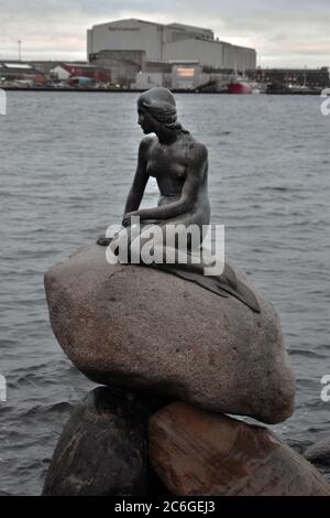 Die kleine Meerjungfrau Statue im Hafen von Kopenhagen von der Langelinie Promenade. Die Fabriken und die industrielle Umgebung sind deutlich hinter der Meerjungfrau zu sehen. Stockfoto