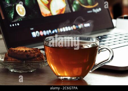 Glasbecher mit Tee und Keksen auf einem Schreibtisch mit Laptop Tee Pause von geliebten Job. Arbeiten Sie online oder offline oder im Büro, freiberuflicher Job Stockfoto