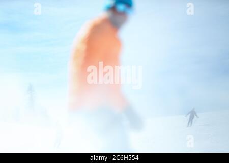 Konzeptuelle Fotografie. Ein männlicher Skifahrer in einer orangen Jacke ist im Vordergrund unscharf. Im Hintergrund fährt ein Skifahrer Stockfoto