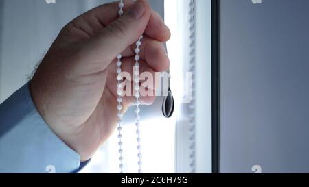 Bild mit der Hand eines Mannes in einem Hotelzimmer, Schlafzimmer oder Büro, Öffnen eines vertikalen Vorhangs am Fenster Stockfoto