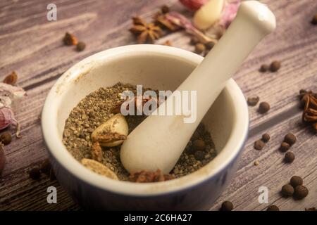 Piment in einem Keramikmörtel und verstreute Knoblauchzehen, Sternanis und Piment auf einem Holztisch. Nahaufnahme Stockfoto