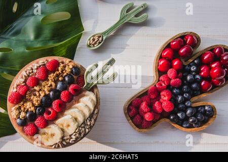 Acai Smoothie, Müsli, Samen, frisches Obst in einer Holzschale mit Kaktuslöffel. Teller gefüllt mit Beeren auf weißem Holzhintergrund Stockfoto