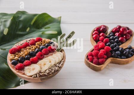 Acai Smoothie, Müsli, Samen, frisches Obst in einer Holzschale mit Kaktuslöffel. Teller gefüllt mit Beeren auf weißem Holzhintergrund Stockfoto