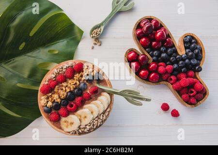 Acai Smoothie, Müsli, Samen, frisches Obst in einer Holzschale mit Kaktuslöffel. Teller gefüllt mit Beeren auf weißem Holzhintergrund Stockfoto