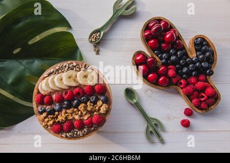 Acai Smoothie, Müsli, Samen, frisches Obst in einer Holzschale mit Kaktuslöffel. Teller gefüllt mit Beeren auf weißem Holzhintergrund Stockfoto