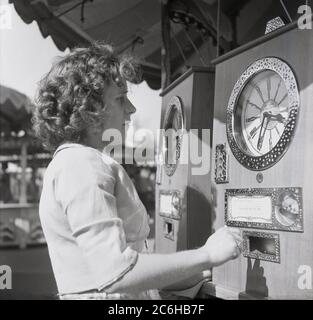 1960er Jahre, historisch, draußen auf einem Jahrmarkt, eine junge Frau, die ein Penny-Arcade-Spiel spielt, ein Uhrspiel mit der Aufschrift „Big Hand on Red to Win“. Beim Spielen wird die Uhr durch Drehen des Knopfes nach rechts aufgewickelt. Entwickelt und hergestellt vom ehemaligen Luftschiffsmechaniker William Bryan aus Derby, England, Großbritannien, und urginell patentiert im Jahr 1930, wurde das Uhrenamusement-Spiel zu einem der erfolgreichsten unter den vielen, die er erfand. Stockfoto