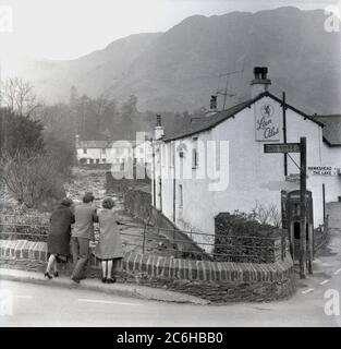 1950er Jahre, historisch, Coniston, Lake District, drei Besucher stehen mit Blick auf einen Bach, Church Beck, neben dem Black Bull Inn, einem alten postkutschenlokal und Hotel, mit einem Lions Ales-Schild, wahrscheinlich nicht traditionelle Seebad-Wanderer, da die beiden Damen Schuhe mit Absätzen tragen. Ein Straßenschild für Hawkshead und den See. Im Hintergrund ein Berg, der als „Alter Mann“ bekannt ist. Stockfoto