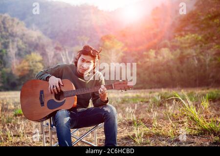 asiatischer jüngerer Mann und akustische Gitarre sitzen auf offenem Feld gegen schöne Sonne Licht Stockfoto
