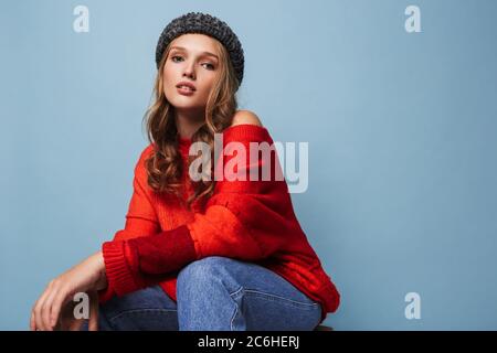 Junge schöne Dame mit welligen Haaren in Hut und roten Pullover traumhaft in der Kamera über blauen Hintergrund Stockfoto