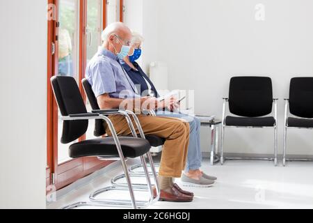 Seniorenpaar sitzt mit Gesichtsmasken in einem hellen Warteraum eines Krankenhauses oder eines Büros - der Mann liest ein Testergebnis - die Frau schaut auf ein s Stockfoto