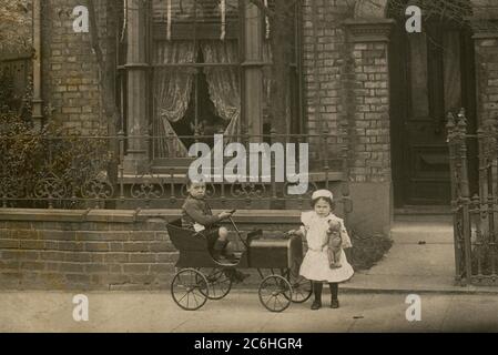 England. Ungefähr 1910. Ein Junge und seine Schwester posieren für ein Foto vor ihrem Haus. Der Junge sitzt in einem Tretwagen und hat einen Spielzeugbus-Schaffner-Automaten über seiner Schulter. Das Mädchen hält einen Teddybär in der Hand. Stockfoto