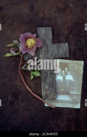 Verwittertes Holzkreuz auf angetünchten Metall mit verblassten Lentonrose oder Helleborus orientalis und Foto der 20er Jahre Familiengruppe Stockfoto
