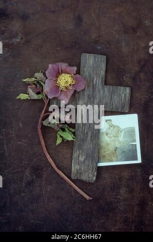 Verwittertes Holzkreuz auf angetünchten Metall mit verblassten Lentonrose oder Helleborus orientalis und Foto von 1940er-Mann in Arbeitskleidung im Freien Stockfoto