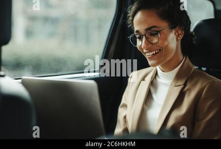 Lächelnde Geschäftsfrau, die mit einem Laptop arbeitet, während sie in einem Taxi reist. Frau mit Laptop auf dem Rücksitz des Autos. Stockfoto