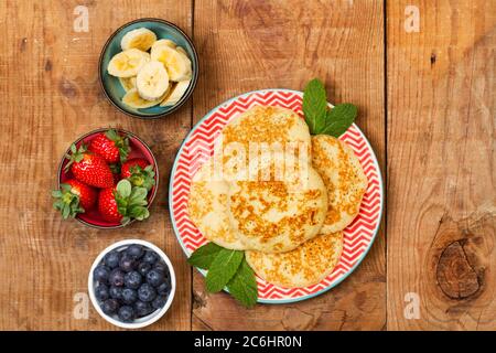 Pfannkuchen auf einem roten Teller und Früchte in Schüsseln auf einem Holztisch in der Draufsicht Stockfoto