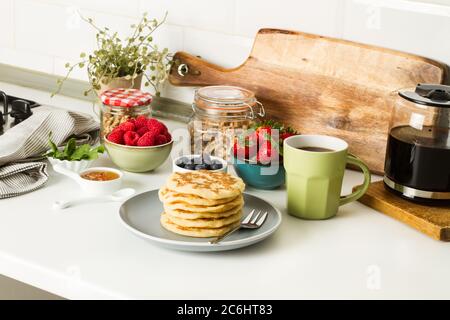 Pfannkuchen mit Beeren und Honig auf einem grauen Teller und mit einer Tasse Kaffee auf einer Küchentheke Stockfoto