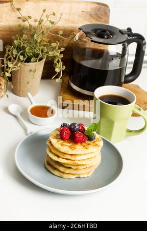 Pfannkuchen mit Beeren und Honig auf einem grauen Teller und mit einer Tasse Kaffee auf einer Küchentheke Stockfoto