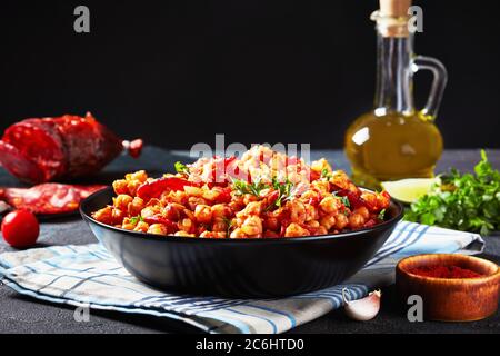 Nahaufnahme von Garbanzos fritos, heißem Chickpea Eintopf mit in Scheiben geschnittenen Chorizo, Schinken, Tomaten und Gewürzen in einer schwarzen Schüssel auf einem Betontisch mit Zutaten an Stockfoto
