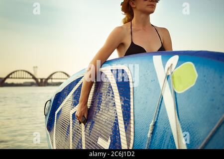 Zeit zum Genießen. Junge attraktive Frau trägt Paddle Board, SUP. Aktives Leben, Sport, Freizeitangebot. Kaukasische Frau auf Reise Bord im Sommer Abendzeit. Urlaub, Resort, Genuss. Stockfoto