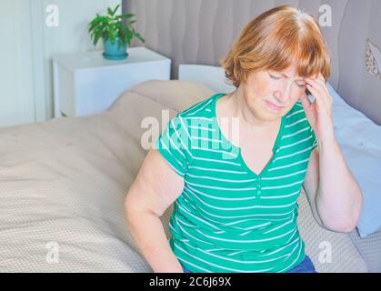 Ältere Frau mit Kopfschmerzen sitzend auf dem Bett zu Hause in Freizeitkleidung. Sie hält ihre Hand an ihre Stirn Stockfoto