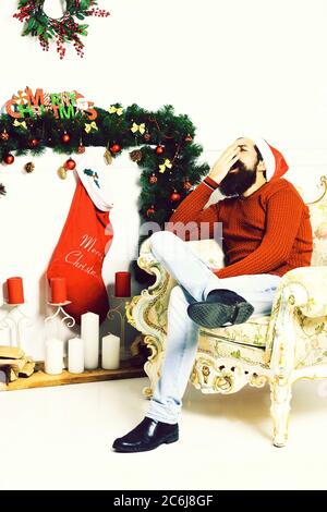 Gut aussehender bärtiger Mann mit langem Bart in Christmas Red Hat und Pullover sitzen und Gähnen auf beige Vintage Sessel um Weihnachten Dekoration, auf weissem Studiohintergrund Stockfoto
