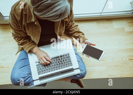 Ältere Frau sieht auf schwarzen Bildschirm des Mobiltelefons arbeiten mit Laptop-Computer. Geschäftsfrau sitzend Schneidermode mit gekreuzten Beinen und gelehnt Stockfoto