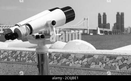 Fernrohr auf rotierender Basis montiert auf einem touristischen Aussichtspunkt im Freien mit verschwommener Stadtlandschaft auf einem Hintergrund. Schwarzweiß-Foto Stockfoto