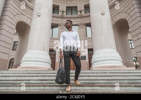 Schöne Eleganz stilvoll gekleideter junger schwarzer Mann mit kabellosen Ohrhörern und Ledertasche in der Hand. Stockfoto