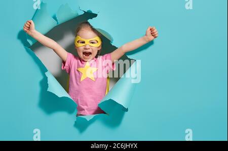 Kleines Kind spielt Superheld. Kind auf dem Hintergrund der hellblauen Wand. Mädchen Power Konzept. Gelbe, rosa und türkisfarbene Farben. Stockfoto