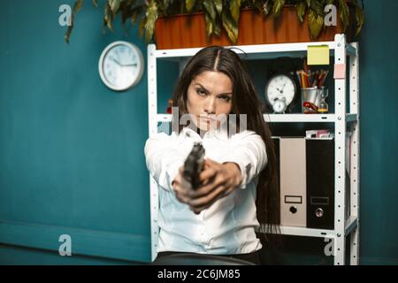 Gefährliche Frau mit Waffe im Büro. Verrückte oder böse junge Geschäftsfrau, die direkt auf die Kamera zielt. Krisenkonzept. Getöntes Bild Stockfoto