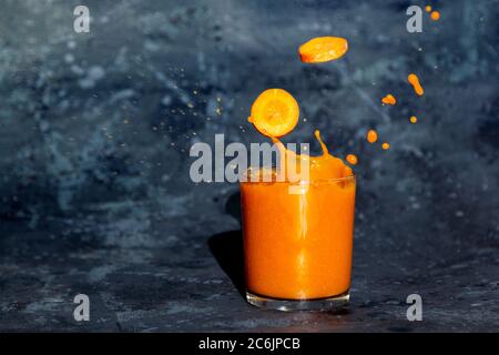 Frische Karottenscheiben fallen in ein Glas mit Karottensaft. Spritzer in Glas auf dunklem Hintergrund Stockfoto