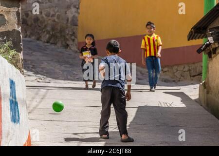 Guatemala, Solola Department, Santa Cruz la Laguna, zwei junge Jungs spielen Fußball oder Fußball auf der Straße, während ein Mädchen in traditioneller Kleidung vorbei geht. Stockfoto