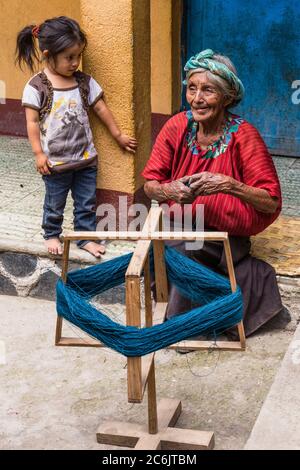 Guatemala, Solola Department, Santa Cruz la Laguna, eine ältere Cakchiquel Maya Frau in traditionellem Kleid Winde Faden auf einem Rahmen, um für das Weben auf einem Webstuhl vorzubereiten, wie ein kleines Mädchen Uhren. Stockfoto