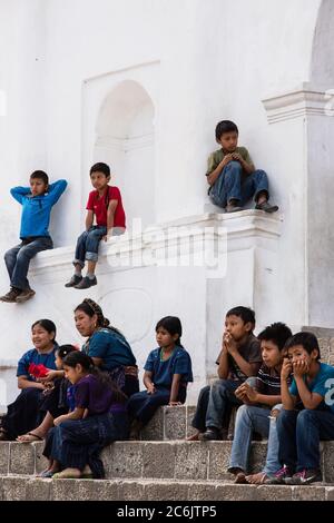 Guatemala, Solola Department, San Antonio Palopo, EINE Cakchiquel Maya-Frau und Kinder beobachten ein Programm von den Stufen der Kirche aus. Die Frau und die Mädchen tragen das traditionelle Kleid von San Antonio Palopo Guatemala, einschließlich der aufwendigen cinta oder Haarwickel, gewebte blaue Huipil Bluse, faja oder Gürtel und corte Rock. Die Jungen tragen alle moderne Kleidung. Stockfoto