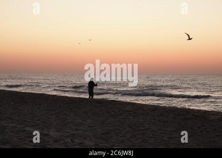 Fischer fängt Fische vom Ufer bei Sonnenuntergang. Der alte Mann hält die Angelrute in den Händen, während er an der Küste steht. Dunkle männliche Silhouette in der Nähe Stockfoto